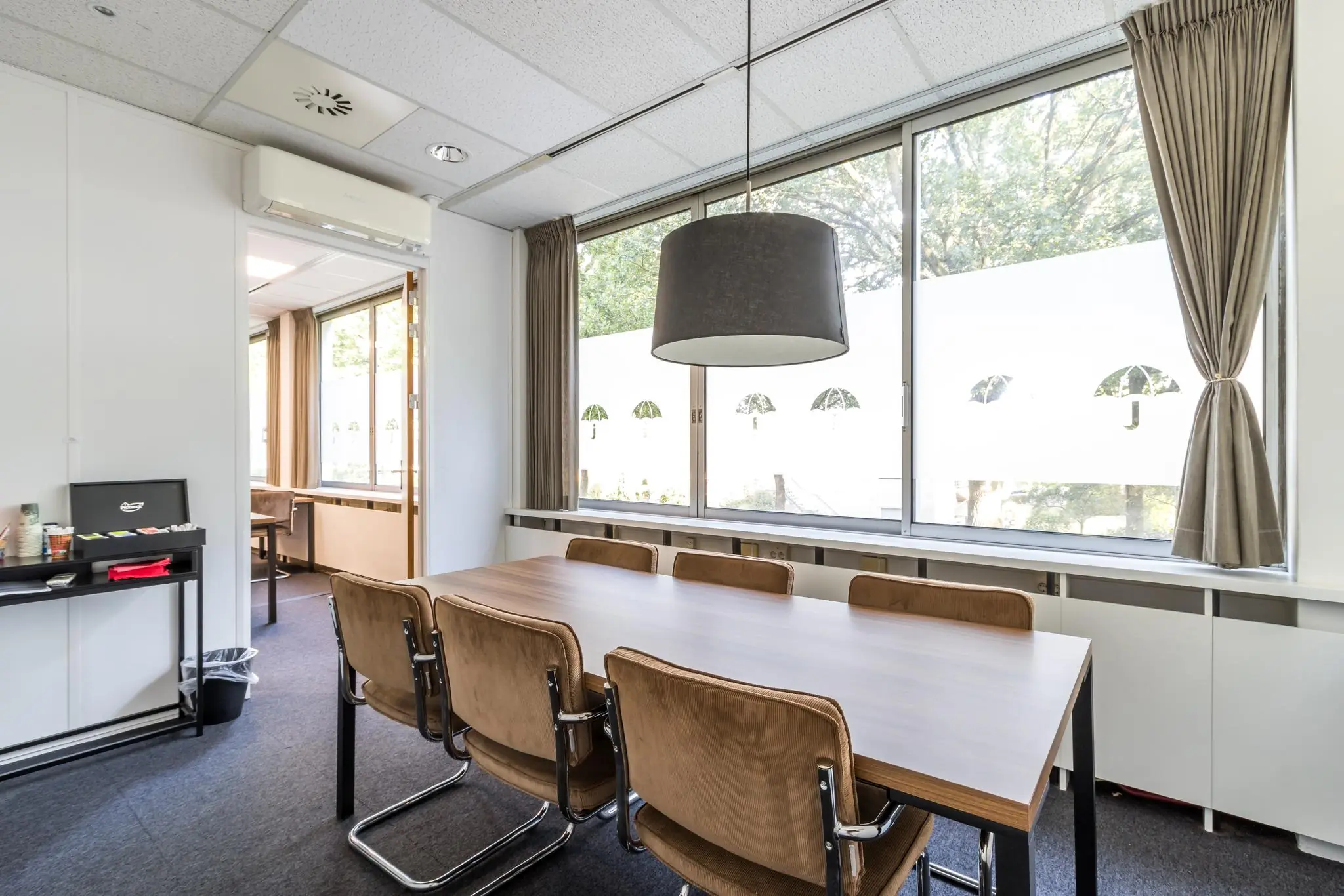 Vergaderruimte aan de Jan van Eycklaan met houten tafel, zes stoelen en grote ramen met uitzicht op groen.